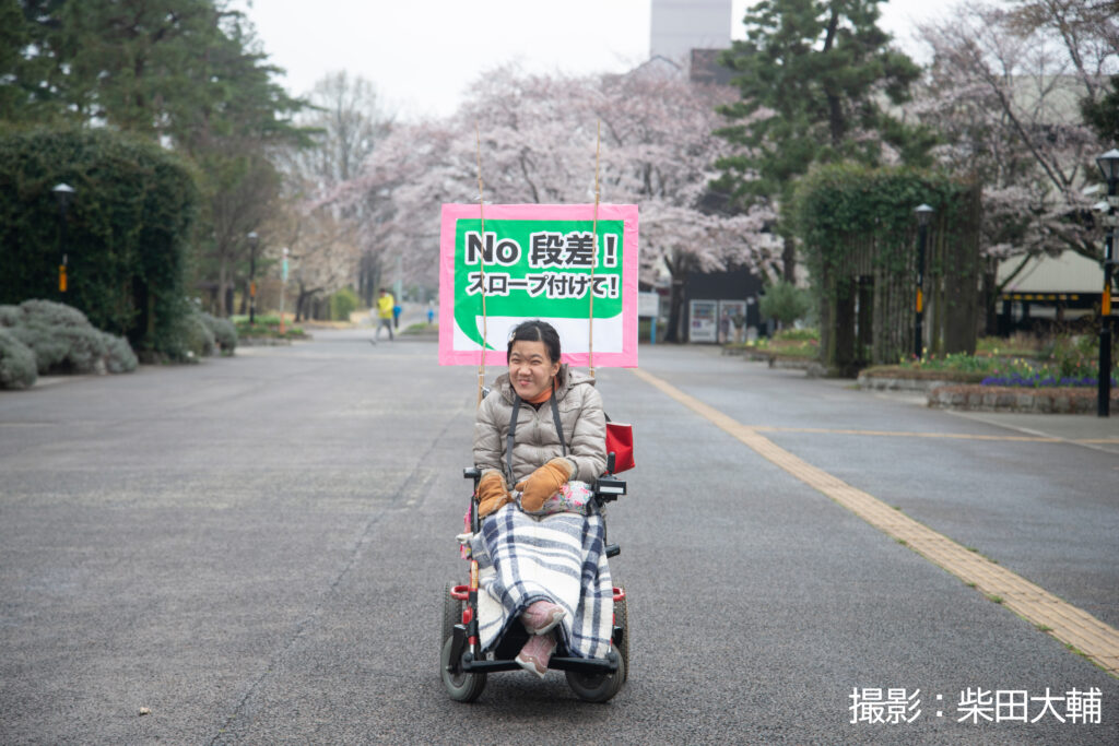 女性の車椅子のパレードの参加者が車椅子の後ろに「No、段差！スロープつけて！」と書いたプラカードをつけて笑顔で微笑んでいる。