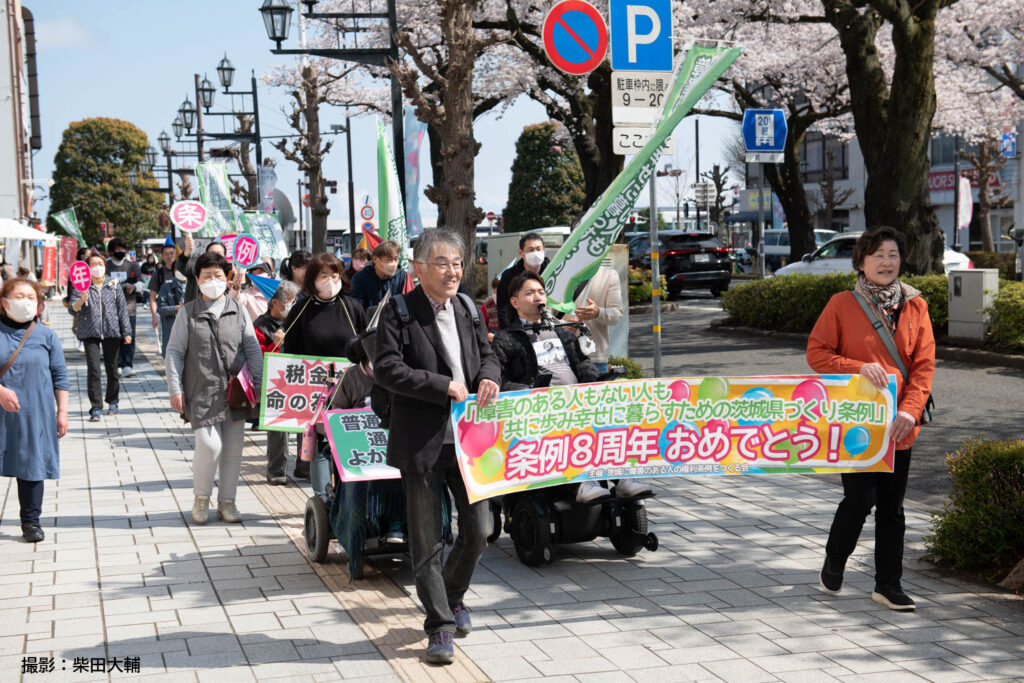 日立市での条例８周年記念パレードの様子。満開の桜のしたを、横断幕をもった参加者がパレードしている。